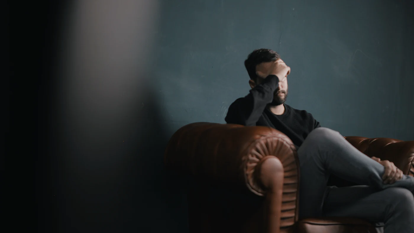 Ein Mann sitzt in einem dunklen Raum auf dem Sofa und stützt den Kopf in die Hand